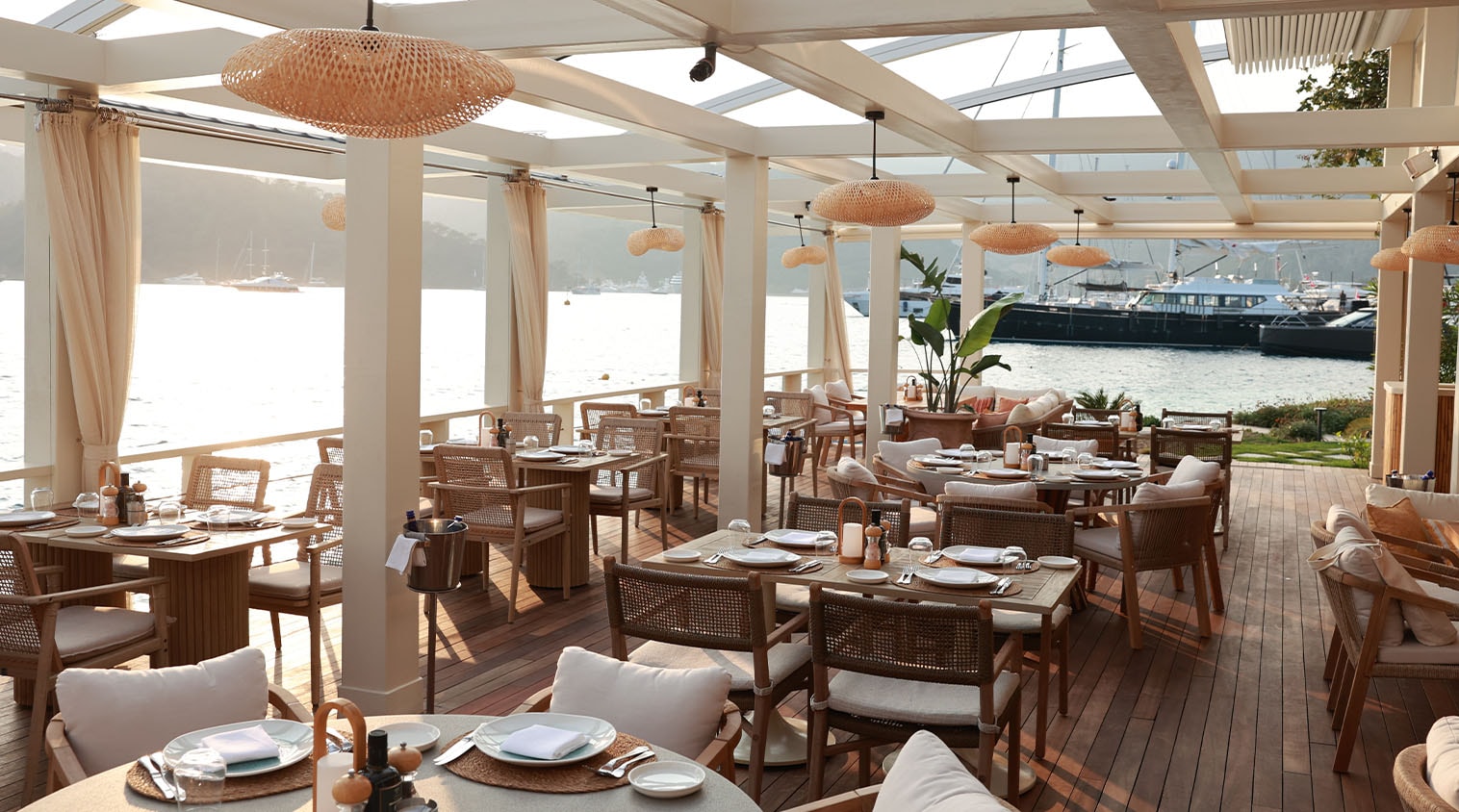 A covered seaside dining terrace with wicker lamps and set tables on a wooden deck at D Breeze