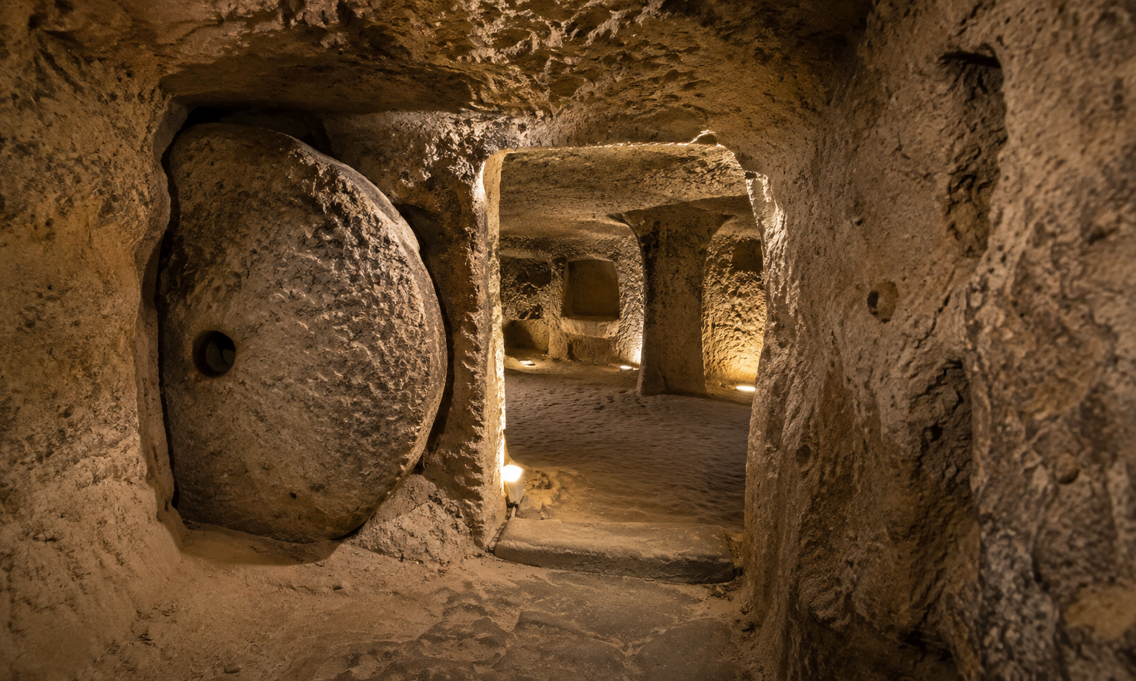 A carved rock passage with a round millstone door opening into a softly lit underground chamber