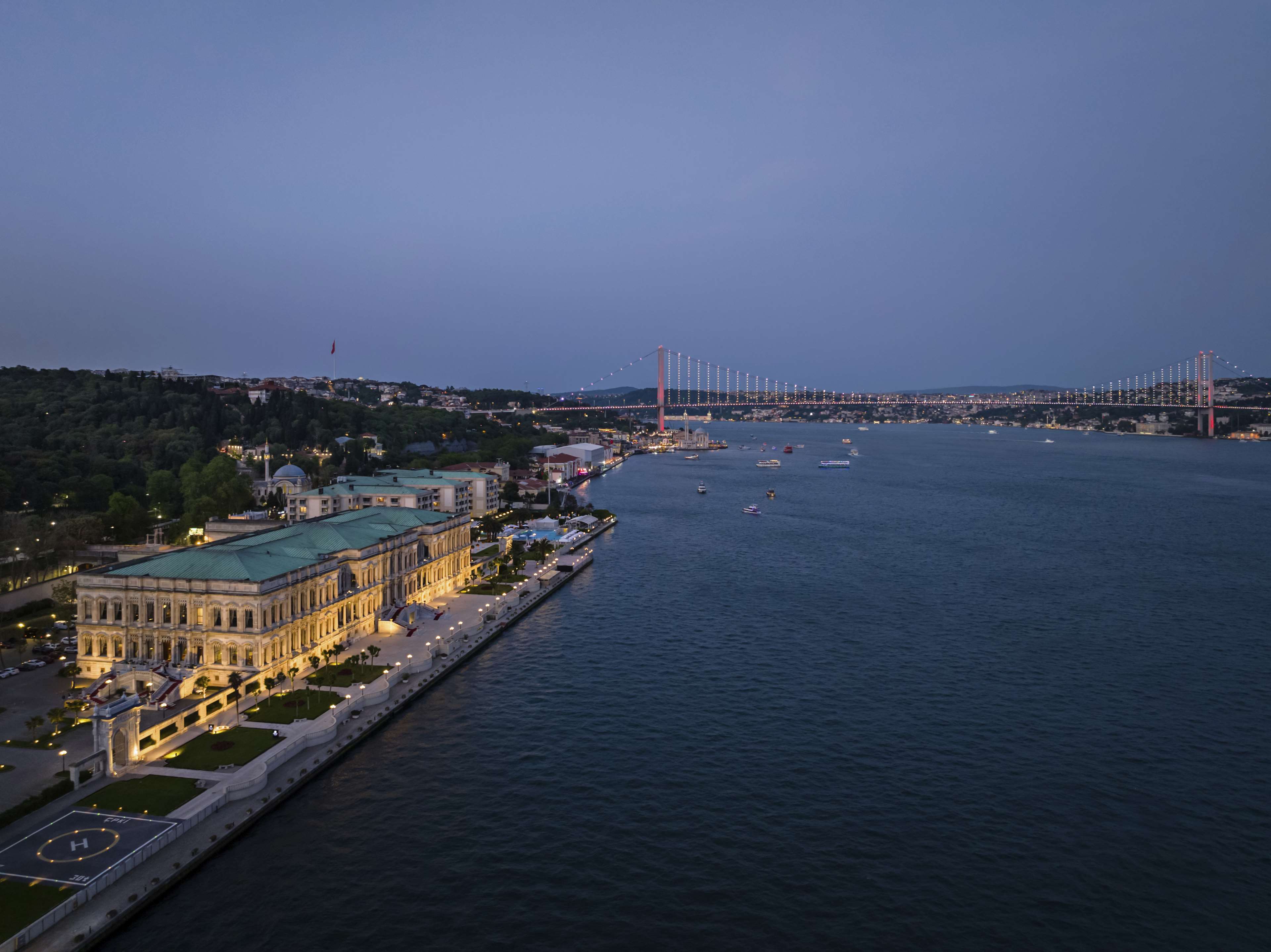Aerial view of Çırağan Palace Kempinski lit at dusk on the Bosphorus, with the bridge beyond