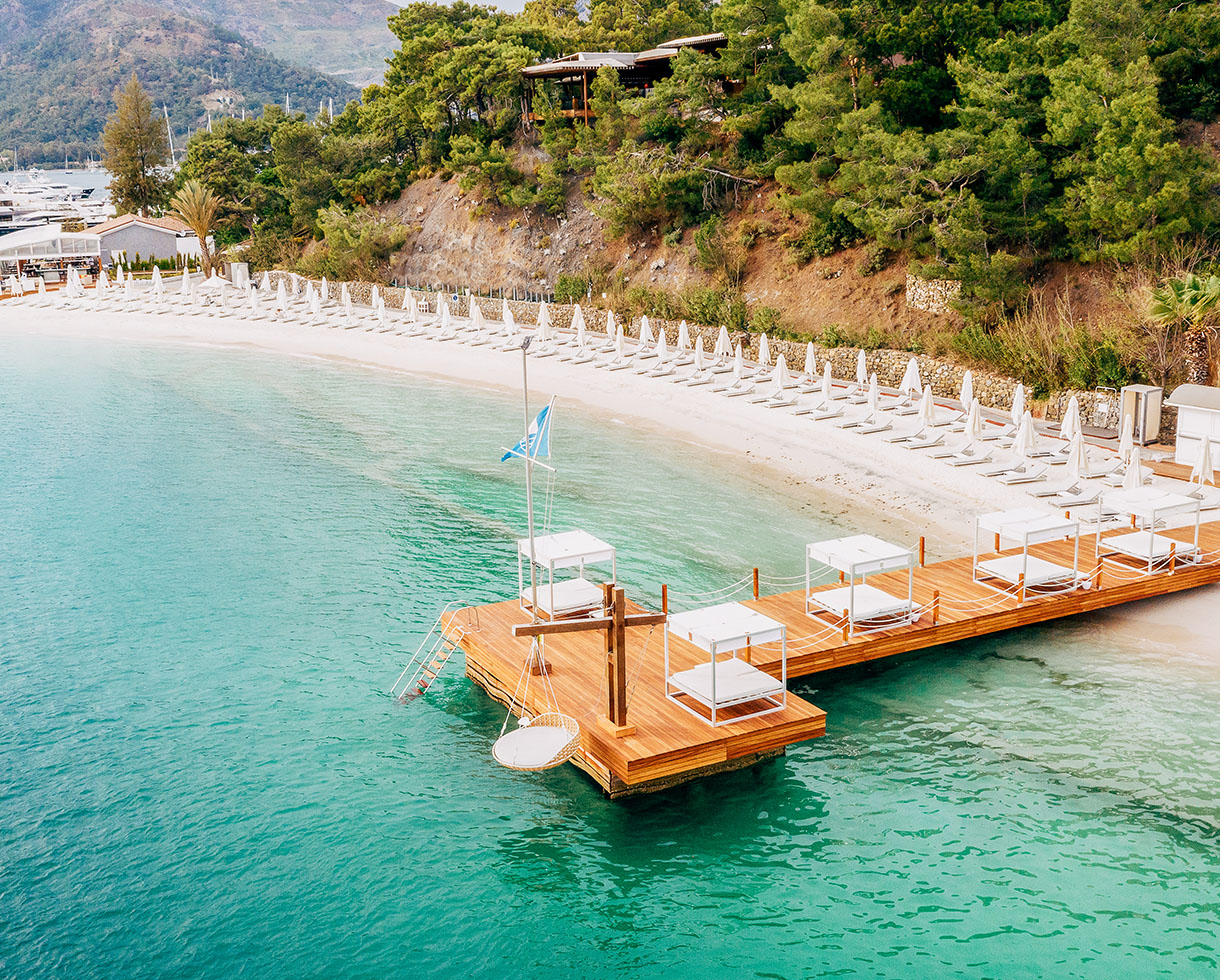 Aerial view of D-Resort Göcek's beach and wooden pier with cabanas over turquoise water
